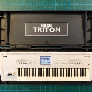 Puede incluir: Un teclado de estación de trabajo/muestreador Korg Triton blanco en un estuche negro. El teclado tiene teclas blancas y negras, una pequeña pantalla y varios botones. La tapa del estuche muestra el logotipo de Korg Triton.