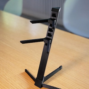 Può includere: Un supporto nero e regolabile per laptop con un supporto verticale e due bracci orizzontali. Il supporto ha una base a tre gambe per la stabilità ed è progettato per sollevare un laptop per un uso ergonomico.