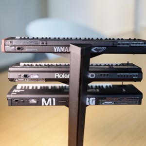 Op de afbeelding: Een zwarte standaard met drie kleine synthesizers. Op de bovenste synthesizer staat het woord "YAMAHA". Op de middelste synthesizer staat het woord "Roland". Op de onderste synthesizer staan de letters "MI".
