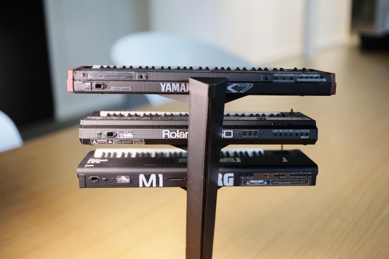 Op de afbeelding: Een zwarte standaard met drie kleine synthesizers. Op de bovenste synthesizer staat het woord "YAMAHA". Op de middelste synthesizer staat het woord "Roland". Op de onderste synthesizer staan de letters "MI".
