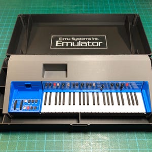 Puede incluir: Un sintetizador gris y azul con teclas blancas, alojado en una caja negra. El sintetizador tiene un panel de control azul con botones rojos y blancos. La tapa de la caja muestra el texto "E-mu Systems Inc. Emulator".