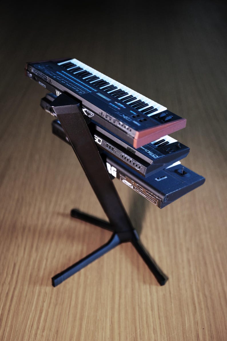 Op de afbeelding: Een zwarte keyboardstandaard ondersteunt drie miniatuursynthesizers. De bovenste synth is gemarkeerd met "Yamaha DX7" met witte toetsen. De middelste synth is gemarkeerd met "Korg DS-80". De onderste synth is gemarkeerd met "Korg".
