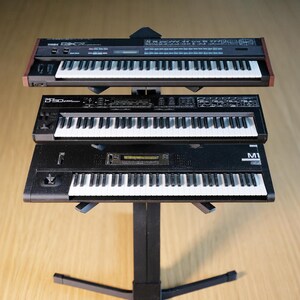 Op de afbeelding: Drie zwarte synthesizers met witte toetsen zijn op een zwarte standaard gestapeld. De bovenste synth heeft een bruine houten rand. De middelste synth heeft de tekst "D-50" erop. De onderste synth heeft de tekst "M1" erop. De standaard heeft een gebogen voet.