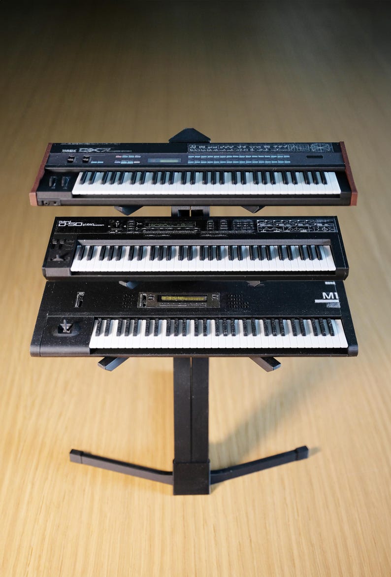 Op de afbeelding: Drie zwarte synthesizers met witte toetsen zijn op een zwarte standaard gestapeld. De bovenste synth heeft een bruine houten rand. De middelste synth heeft de tekst "D-50" erop. De onderste synth heeft de tekst "M1" erop. De standaard heeft een gebogen voet.