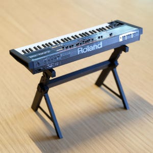 Puede incluir: Un teclado sintetizador Roland Juno-106 en miniatura, negro y gris, con teclas blancas, sobre un soporte negro. El teclado tiene varios botones y deslizadores, con el logotipo de Roland visible. El soporte tiene un diseño simple y angular.