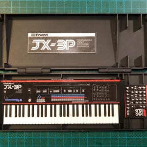 Op de afbeelding: Zwarte Roland JX-3P synthesizer in een open zwarte koffer, met het toetsenbord en bedieningspaneel. De synthesizer heeft een rode streep en de tekst "JX-3P". Een aparte module staat rechts. De koffer ligt op een groene snijmat.