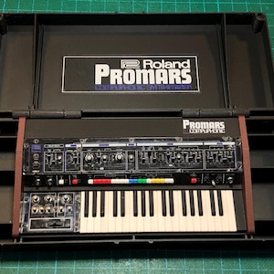 May include: A black Roland Promars Compuphonic Synthesizer in a black case. The synthesizer features a keyboard, knobs, and sliders. The case is open, displaying the instrument. The text "Roland Promars Compuphonic Synthesizer" is visible.