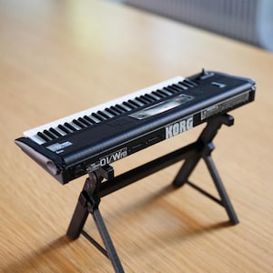 Puede incluir: Un teclado sintetizador Korg 01/Wfd negro con teclas blancas y negras, apoyado en un soporte negro. El teclado tiene el logotipo de Korg y otro texto. El soporte no está incluido.