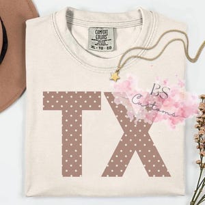 Peut inclure: Un t-shirt à manches courtes de couleur crème arborant les lettres TX en marron avec un motif à pois blancs. Le t-shirt présente une étiquette de marque Comfort Colors à l'encolure intérieure et un collier pendentif étoile dorée.