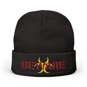 Op de afbeelding: Zwarte beanie met het woord "BEWARE" in rode, getextureerde letters en een geel biohazard-symbool. De beanie is gemaakt van een zacht materiaal en heeft een omgeslagen manchet.