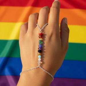 Può includere: Una catena da mano color argento con una fila verticale di perline multicolori in rosso, arancione, giallo, verde, blu, viola, nero, oro, azzurro, bianco e cristallo trasparente, collegate da una sottile catena a un braccialetto.