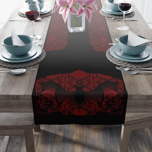 May include: Black table runner with a red bat design. The runner features a detailed bat silhouette with intricate lace-like patterns. The background is black, and the bat is red. The runner is on a wooden table with place settings.