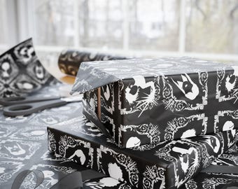 May include: Two wrapped presents, covered in black paper with a white decorative design, are stacked on a table. A roll of matching paper, scissors, and a ribbon are also visible, suggesting gift wrapping.
