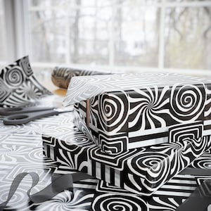 May include: Three gift boxes wrapped in black and white patterned paper with swirling and geometric designs. The boxes are stacked on a table with matching wrapping paper, scissors, and a ribbon. The overall theme is gift wrapping.