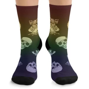 Puede incluir: Un par de calcetines con un fondo degradado de marrón, verde y azul. Los calcetines presentan un patrón repetido de calaveras blancas y diseños de insectos. Los dedos de los pies y los talones son negros.