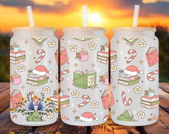 Op de afbeelding: Drie glazen bekers met houten deksels en rietjes, versierd met kerstillustraties. De ontwerpen omvatten boeken, kaarsen, zuurstokken en kerstmutsen in pastelkleuren. De bekers staan op een houten oppervlak tegen een zonsondergang.