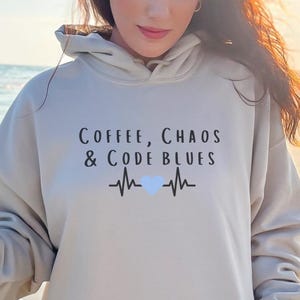 Könnte beinhalten: Ein beigefarbener Kapuzenpullover mit dem Text "Coffee, Chaos & Code Blues" und einer Herzschlag-Grafik. Eine gelbe isolierte Wasserflasche wird in der Hand gehalten. Das Bild wird an einem Strand mit dem Meer im Hintergrund aufgenommen.