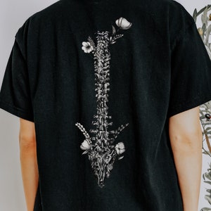 May include: Black t-shirt featuring a detailed grayscale illustration of a spine adorned with floral elements. The design is centered on the back of the shirt. The shirt has short sleeves and a relaxed fit.