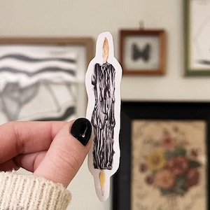 May include: A sticker featuring a black candle with a yellow flame. The candle has a dripped wax design. The sticker is held in front of a wall with framed artwork.
