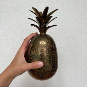 Puede incluir: Un recipiente metálico en forma de piña de color bronce, con una superficie texturizada de patrón de diamantes y una parte superior puntiaguda con hojas. El objeto presenta una costura horizontal en el medio, indicando una tapa, con un acabado pulido.