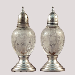May include: A pair of antique salt and pepper shakers. Each shaker features a clear, etched glass body with floral designs and a silver-colored metal top and base. The tops have small holes for dispensing.