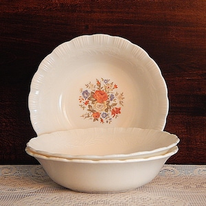May include: Two off-white ceramic bowls with a floral design. The bowls have a scalloped edge and a central bouquet of orange, red, blue, and white flowers. The bowls are stacked on a white lace tablecloth.