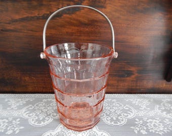May include: A vintage pink glass bucket with a silver handle. The bucket has a geometric design and is likely used for ice or as a decorative piece. The handle is arched and attached to the bucket's rim.