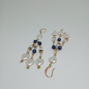 May include: Dangling earrings featuring white heart-shaped beads, navy blue and white round beads, and gold-colored accents. The earrings have a gold-colored hook closure and are displayed against a white background.