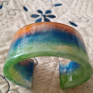 May include: A colorful cuff bracelet with a vibrant, blended design. The bracelet features a gradient of colors, including orange, blue, and green. The open-ended design allows for easy wear. The bracelet is a unique accessory.