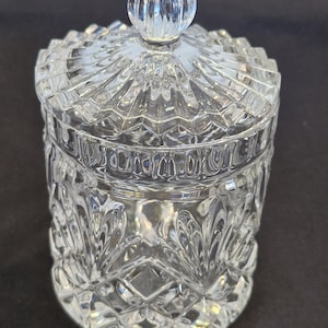 May include: A clear glass jar with a matching lid featuring a spherical knob handle. The body and lid are decorated with a pressed geometric pattern consisting of diamond shapes and vertical fan-like motifs, creating a textured, faceted appearance.