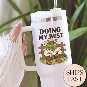 May include: White tumbler with a handle and a lid, featuring a cartoon frog in a cowboy hat and the text "DOING MY BEST." The frog is sitting on a fence. The tumbler has a straw and is held by a person wearing a white sweater.