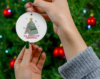 May include: A white round Christmas ornament with a green and brown book stack shaped like a Christmas tree. The ornament has the text "All Booked for Christmas" on it.