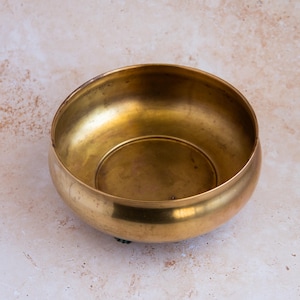 May include: A round, antique brass bowl with a wide rim and a slightly curved base. The bowl has a polished, golden finish and sits on a neutral-toned surface. The interior of the bowl is smooth and reflective. This bowl is suitable for decorative or functional use.