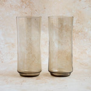 May include: Two tall, cylindrical drinking glasses. The glasses are made of clear, tinted glass with a light brown hue. Each glass has a slightly flared base and a straight, smooth body. The glasses are empty and sit on a neutral-toned surface.