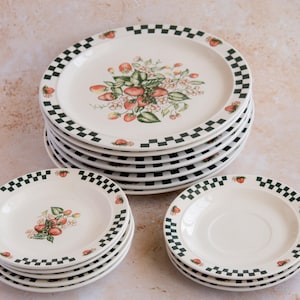 May include: A collection of white ceramic plates featuring a strawberry motif and a green and black chequered border. The plates are stacked in three groups, demonstrating a dinnerware set, ideal for serving food.