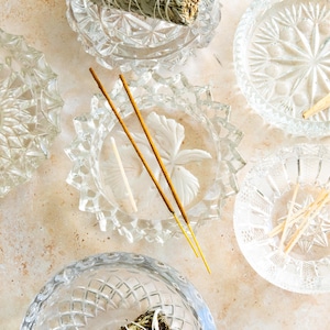 May include: Overhead shot of several clear glass ashtrays with decorative patterns. Some ashtrays contain sage bundles, incense sticks, and matches. The arrangement is on a light-colored, textured surface.