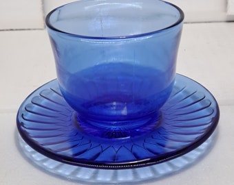 May include: A cobalt blue glass teacup and saucer set. The teacup is a rounded cylinder shape, and the saucer has a ridged, fan-like design. The set is made of transparent glass, and the color is consistent throughout.