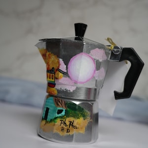 May include: A silver stovetop espresso maker with a black handle and lid. The body is hand-painted with colorful landscape scenes, including a house and a trolley. The text "Pho. Pho. R.D" is visible on the base.