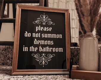 May include: A framed sign with a black background and gold frame. The sign reads "Please do not summon demons in the bathroom". The sign is sitting on a granite countertop with dried pampas grass in vases.