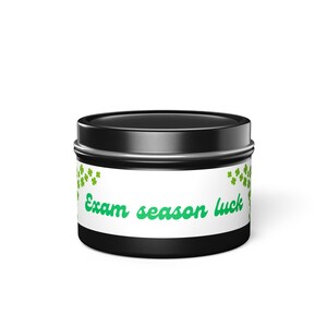 Puede incluir: Un tarro de vela negro con una etiqueta blanca con el texto "Exam season luck" en escritura verde. Los diseños de tréboles verdes están en la etiqueta. El tarro tiene una tapa y una base negras.