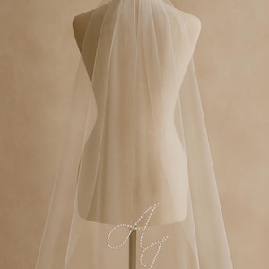 May include: A sheer, ivory-colored wedding veil is draped over a mannequin. The veil features a delicate pearl embellishment in the shape of the letters "A" and "J". The veil is long and flowing, with a soft, ethereal appearance.