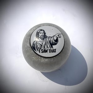 Può includere: Un oggetto decorativo ovale grigio e glitterato. Presenta un'immagine circolare di Gesù che indica, con il testo "I SAW THAT" sotto. L'oggetto è probabilmente un gadget o un regalo scherzoso.