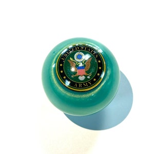 May include: A green and gold round gear shift knob with a United States Army emblem. The emblem features a bald eagle with a shield and stars.