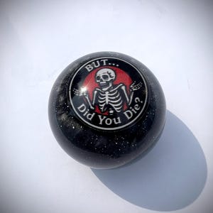 Può includere: Una sfera nera e glitterata con un design circolare con uno scheletro e il testo "BUT... Did You Die?". Il design ha uno sfondo rosso e testo bianco. Probabilmente un oggetto decorativo o un accessorio.