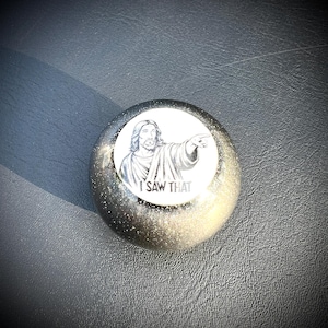 Puede incluir: Objeto decorativo redondo, brillante, plateado y dorado con una imagen circular blanca de Jesús señalando. El texto "I SAW THAT" está debajo. El objeto está sobre una superficie gris oscuro.
