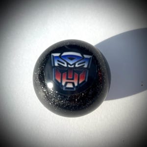 May include: A black, spherical object with a glittery surface. The object features a colorful emblem with blue, red, and white accents. The emblem is a stylized robot face, reminiscent of the Transformers franchise.