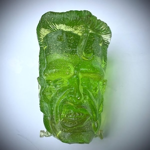 May include: A translucent, lime green sculpture of a face with detailed features, including hair, a prominent nose, and a defined mouth. The sculpture has a textured surface and appears to be made of glass or a similar material. The face has a slight grin.