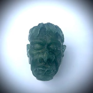 Può includere: Una scultura traslucida color verde acqua raffigurante il volto di un mostro con gli occhi chiusi e la bocca cucita. La scultura ha una superficie strutturata e dettagli visibili, come le orecchie e i capelli. Il volto è centrato su uno sfondo bianco.