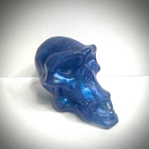 May include: A blue, glittery, resin skull sculpture with a detailed, realistic design. The skull is tilted to the side, with the jaw slightly open.