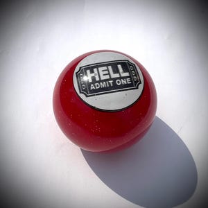 May include: A red, round object with a glossy finish and a white circular emblem. The emblem reads "HELL ADMIT ONE" in black text. The object has a slight glitter effect and casts a shadow.
